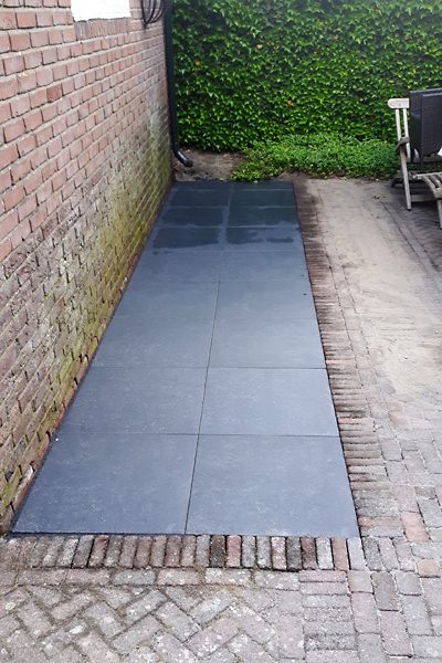 Smal pad van donkere natuursteentegels langs een oude bakstenen muur